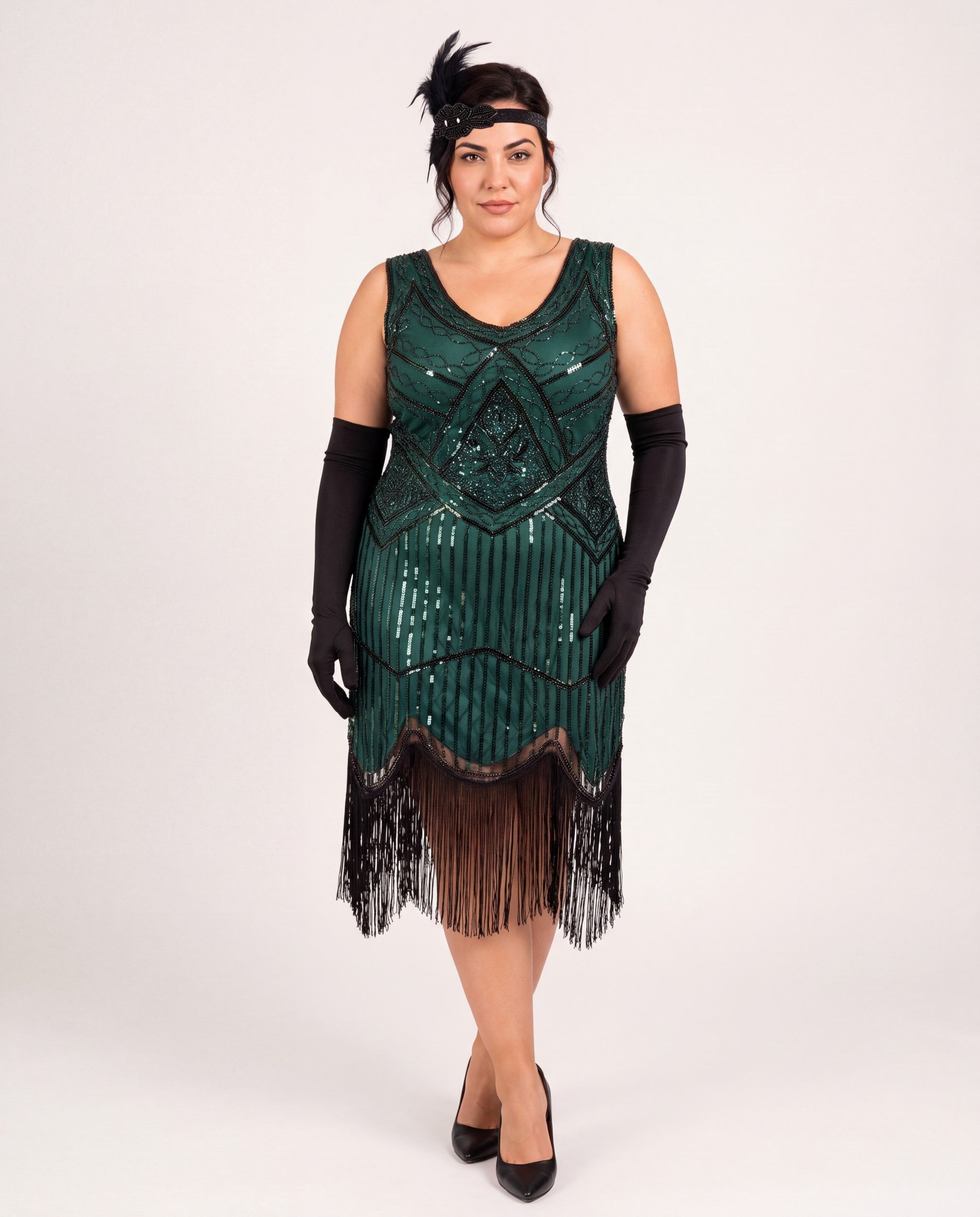 A woman modeling a 1920s-style plus size flapper dress in dark green sequins with black fringe, paired with black gloves, a black headband, and black heels against a plain light gray studio background.