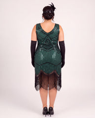 A woman shown from the back wearing a dark green 1920s-style plus size flapper dress with black fringe, black elbow-length gloves, and black heels against a plain light gray studio background.