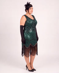 A woman modeling a dark green 1920s-style plus size flapper dress with black fringe, black gloves, a black headband, and black heels against a plain light gray studio background.