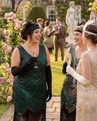 Three women in 1920s flapper-style dresses converse in a garden setting, holding champagne glasses. One dress is dark green with black fringe and beading, another is cream with gold beading, and the background shows other guests and a statue.